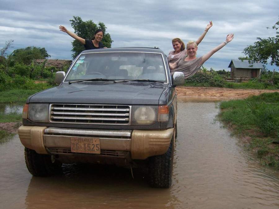 Three women waving from muddy SUV driving through flooded road