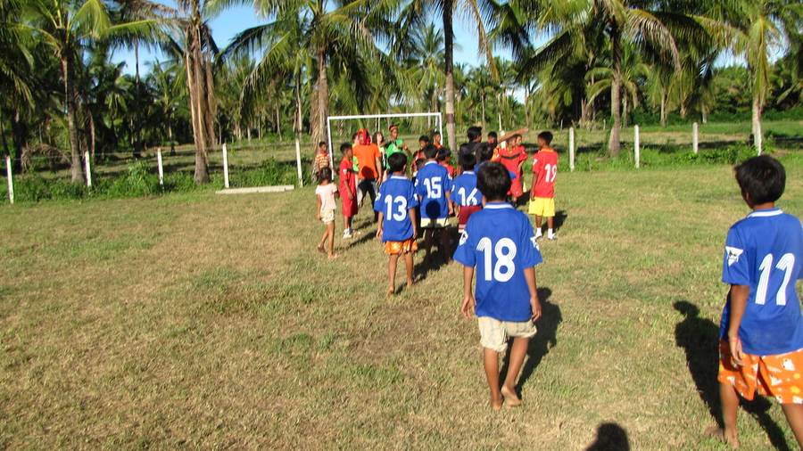 Children in blue numbered jerseys walking toward goal on soccer field with palm trees