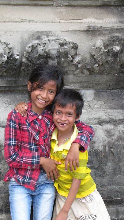 Two children smiling sitting together in front of carved stone wall